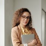 A confident woman in formal attire holding a laptop, standing in an office environment and looking at the camera.